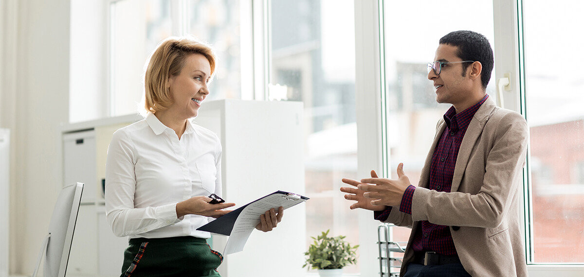 Employer talking to employee while holding a clipboard in an office.